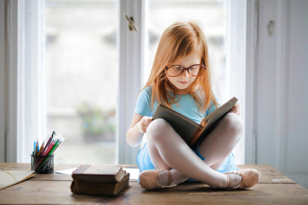 Little girl with red hair and glasses on reading a book while sitting on a wooden table 